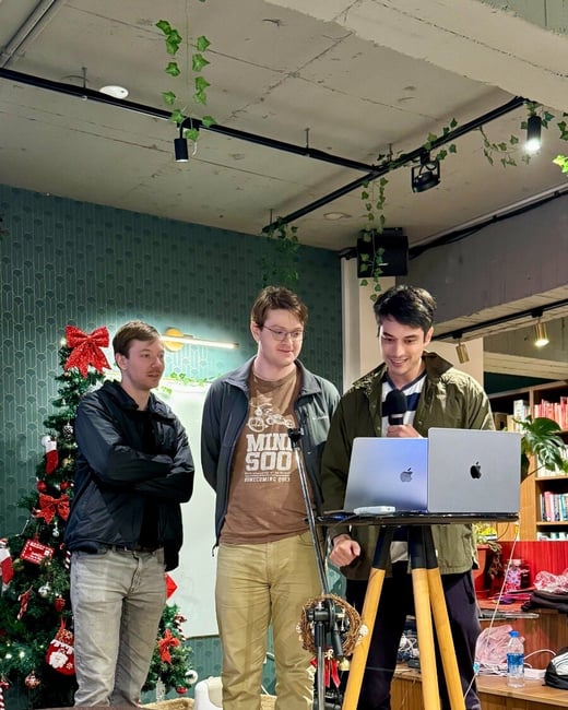 Three men are gathered indoors, possibly for an informal presentation or recording session, with one man speaking into a microphone while standing next to two open laptops set up on a tripod stand. A decorated Christmas tree is visible to the left, suggesting a holiday season setting.