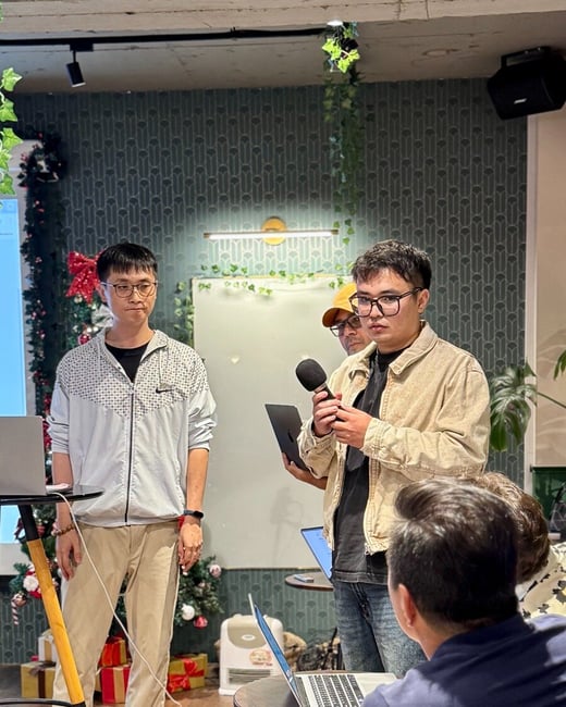 A photograph captures an indoor presentation or meeting where two men are standing near a screen, one speaking into a microphone, set against a backdrop of patterned wallpaper and Christmas decorations.