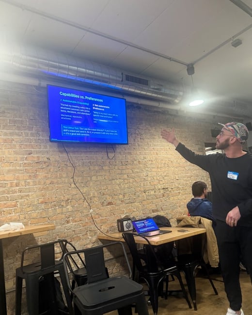 A man in a black shirt and patterned cap is gesturing towards a television screen mounted on a brick wall, which displays a presentation slide about capabilities versus preferences.