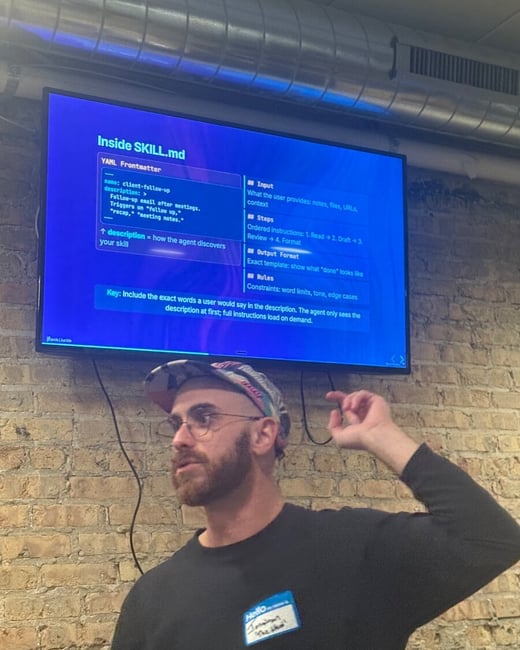 A man wearing a cap and glasses gestures toward a large screen displaying technical documentation for 'Inside SKILL.md' against a brick wall background.