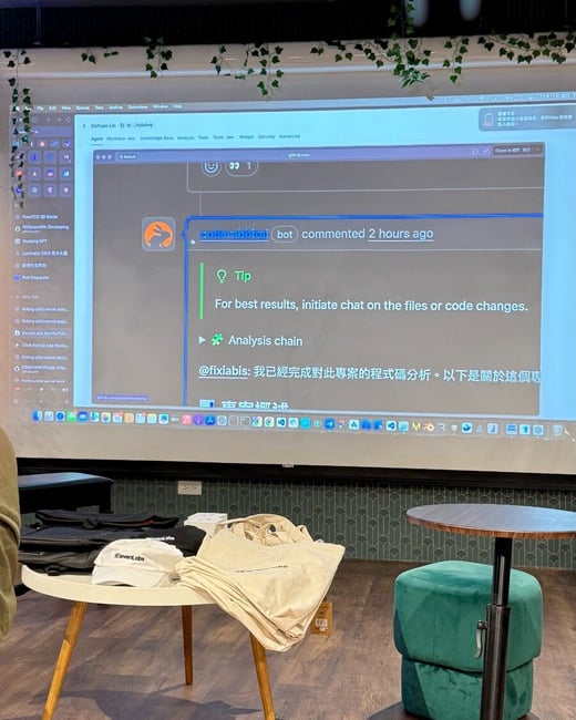 A photograph taken indoors at what appears to be a tech event or presentation, showing a large projected screen displaying a code review interface with comments from a 'coderabbitai' bot, alongside a table holding event merchandise (caps and tote bags).