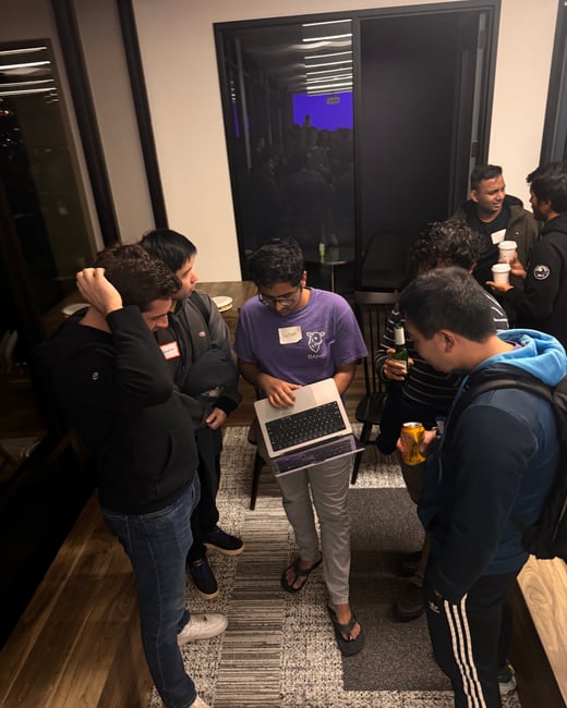A group of young men gathers indoors in low light, standing around one individual who is holding an open laptop, possibly discussing technical details or networking at an event.