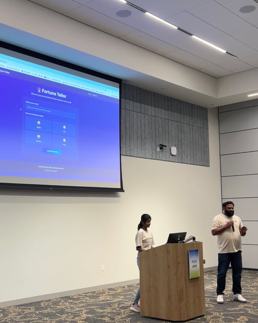 A photograph capturing two presenters demonstrating a web application called "Fortune Teller" on a large projection screen in a modern indoor venue, likely a conference room or library space.