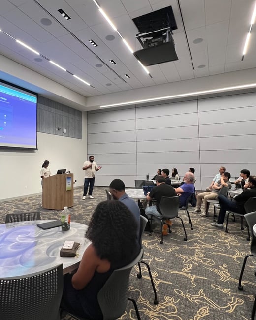 A photograph capturing an indoor presentation or meeting where two people stand near a wooden podium and a large projection screen, addressing an audience seated at tables in a conference room.