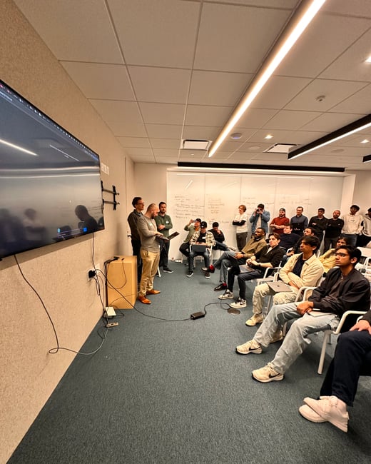 A group of people are gathered in a modern office space for a presentation or demo. One person stands at the front with a laptop, while others sit or stand, watching a large wall-mounted screen.