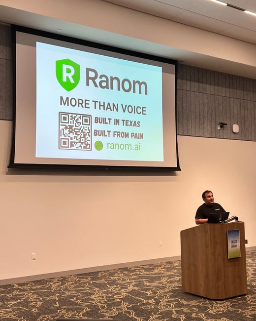 A photograph capturing a man giving a presentation indoors, standing behind a wooden podium, with a large projection screen displaying a slide for the company 'Ranom.'