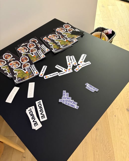 A high-angle photograph showcasing various promotional stickers, including sheets with cartoon illustrations and individual die-cut stickers for brands like PostHog, DRIVE, and AI Tinkerers Chicago, scattered across a black table.