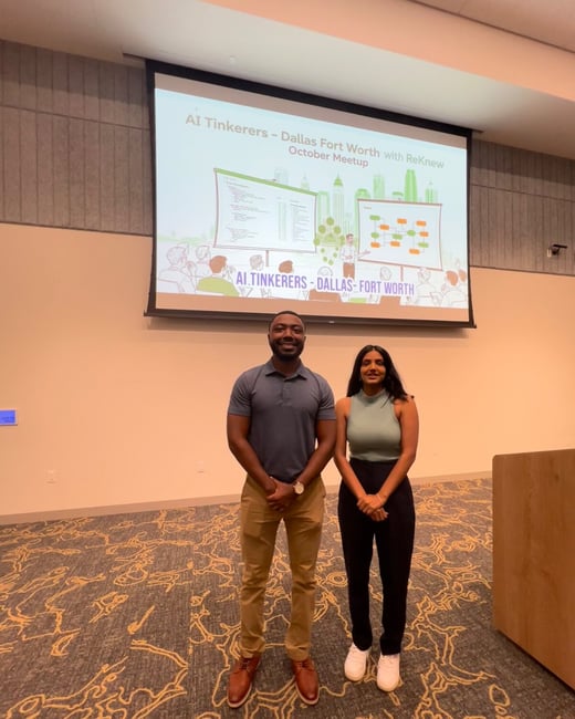 A full-body photograph of a man and a woman standing in an indoor event space, posing in front of a large projection screen displaying information for the 'AI Tinkerers - Dallas Fort Worth' meetup.