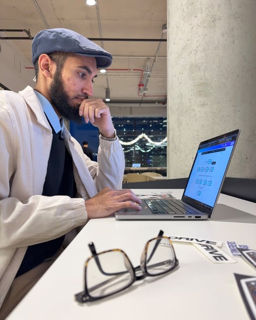 A man wearing a light jacket and a flat cap sits at a white table, intently viewing a flowchart or diagram displayed on his open laptop screen, with his hand resting on his chin in thought. A pair of tortoise-shell reading glasses and some stickers reading "DRIVE" rest on the table in the foreground.