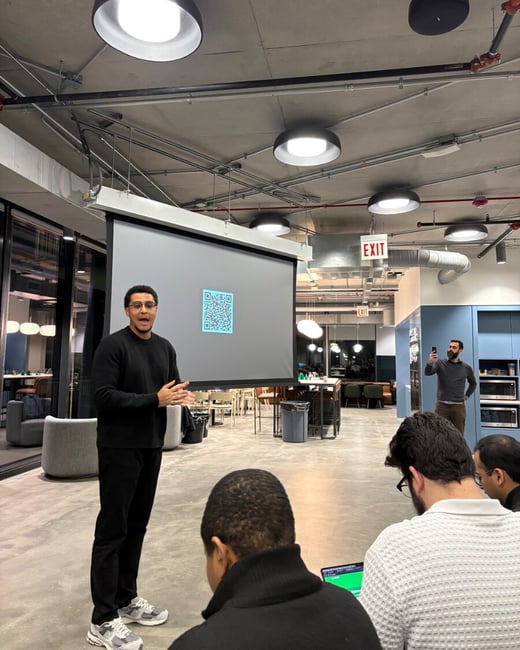 A man in black attire is giving a presentation in a modern office or event space, standing in front of a large screen displaying a bright cyan QR code to a small audience.