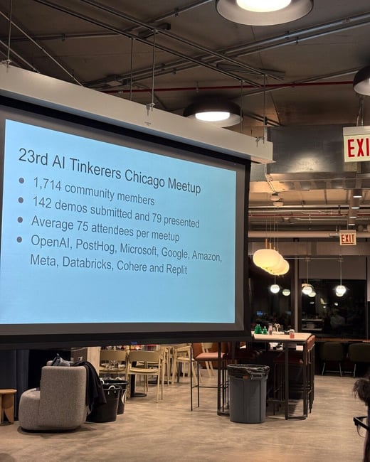 A photograph taken indoors at an event, focusing on a large projection screen displaying bullet points about the 23rd AI Tinkerers Chicago Meetup statistics and sponsors, set against a backdrop of modern industrial architecture and seating.