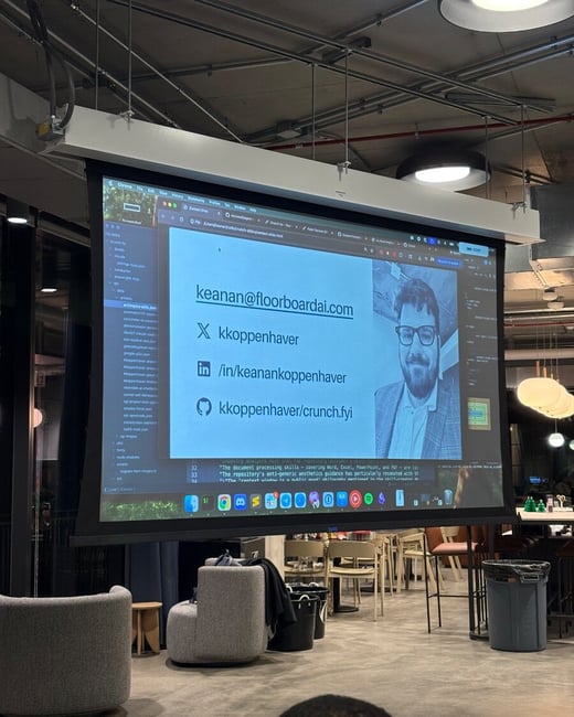 A photograph taken from the audience perspective during an indoor presentation, showing a large projection screen displaying a contact slide with an email, social media handles, and a black and white portrait of a man.