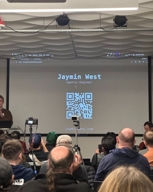 A photograph capturing a live presentation in a room filled with an audience, where a large screen displays the presenter's name, title, and a QR code.