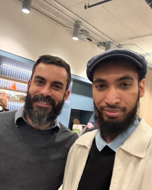 A close-up photograph captures two smiling men with prominent beards posing together indoors, likely in a store or commercial setting with exposed ceiling utilities visible above.