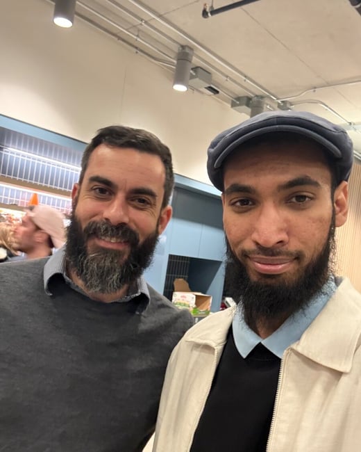 A close-up selfie-style photograph of two men with beards smiling at the camera, taken indoors in a brightly lit commercial or retail environment.