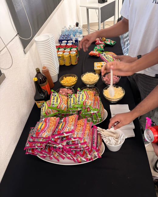 A photograph of a food station at an event, featuring stacks of instant ramen, various toppings like corn and cheese, and drinks on a black tablecloth.