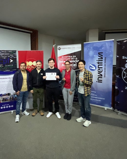 A group of five people posing for a photograph in front of event banners, with the person in the center holding a certificate.