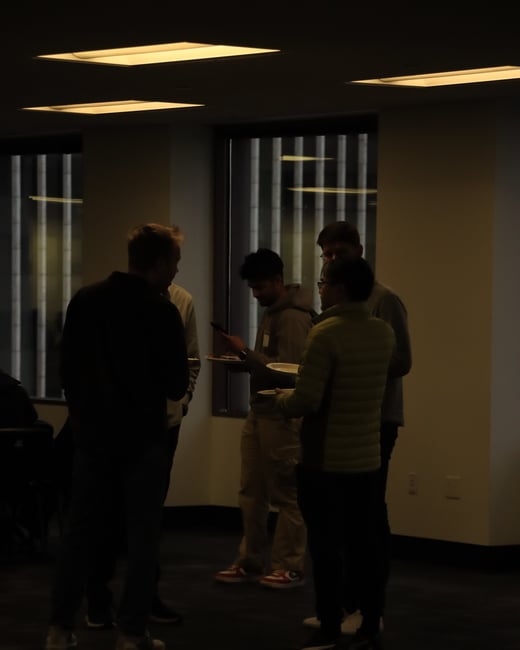 A group of people standing and socializing in a dimly lit indoor space, with some individuals holding plates of food.