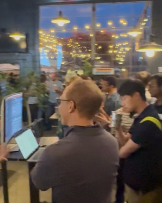 A candid, low-light photograph capturing a group of people gathered indoors at an evening event, where a man in the foreground is observing a monitor displaying code next to an open laptop.