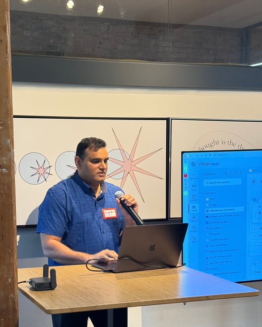 A man in a blue shirt is giving a presentation, standing at a high table with a laptop and holding a microphone while a large screen displays software behind him.