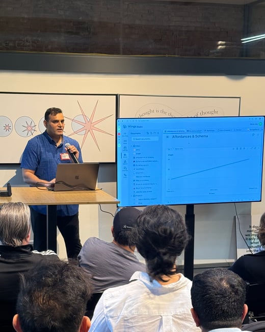 A man is giving a presentation to an audience, standing at a podium with a laptop and a large screen displaying a software interface.
