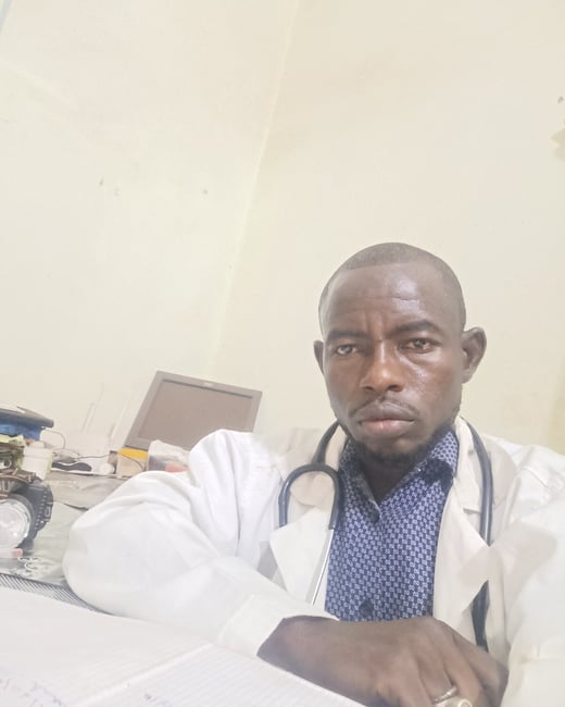 A close-up photograph of a man, likely a doctor or medical professional, wearing a white lab coat and a stethoscope, sitting at a desk indoors and looking directly at the camera.