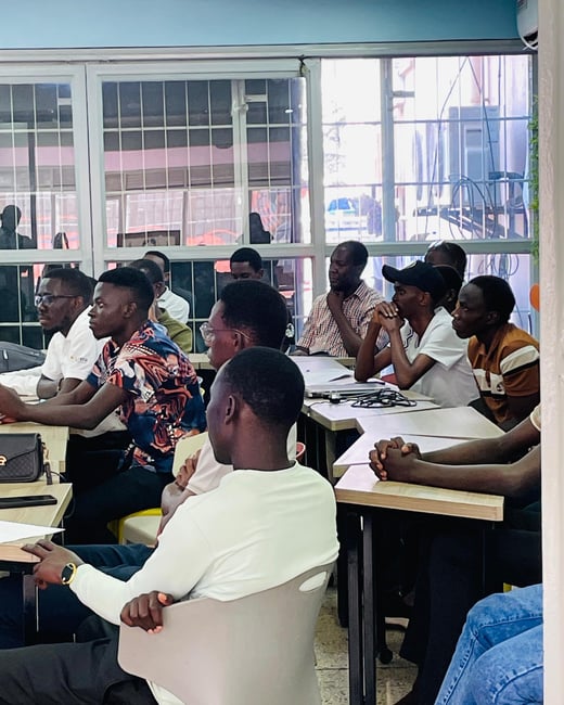 A photograph capturing a group of young men seated at desks in a brightly lit indoor classroom or training room, attending a lecture or presentation. The attendees are facing forward toward an unseen speaker, with large window panes visible in the background.