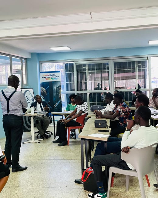 A photograph capturing an indoor workshop or meeting where a man stands addressing a group of approximately ten seated attendees in a brightly lit room with light blue walls.
