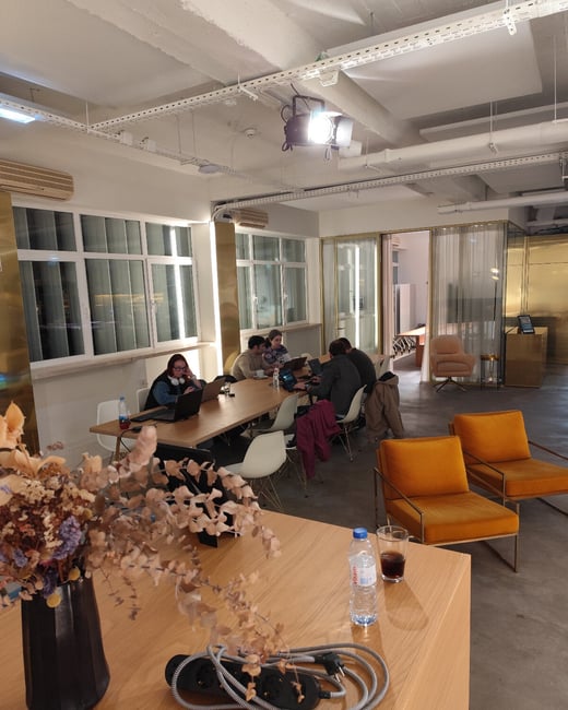 A candid photograph taken indoors showing a modern co-working or office space where five people are seated around a long wooden table working on laptops. The foreground features a close-up of a wooden surface holding a vase of dried flowers, a water bottle, and a coiled power strip.