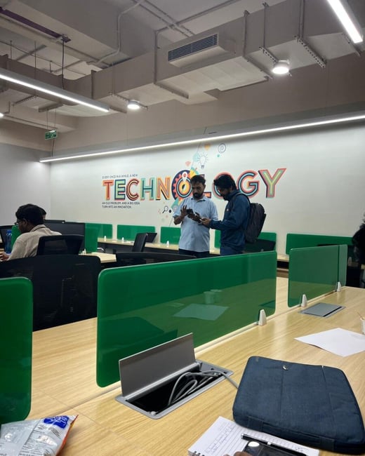 An indoor office workspace featuring wooden desks with green dividers and a large wall graphic that reads 'TECHNOLOGY'. Two men are standing and looking at a phone while another person sits at a desk in the background.