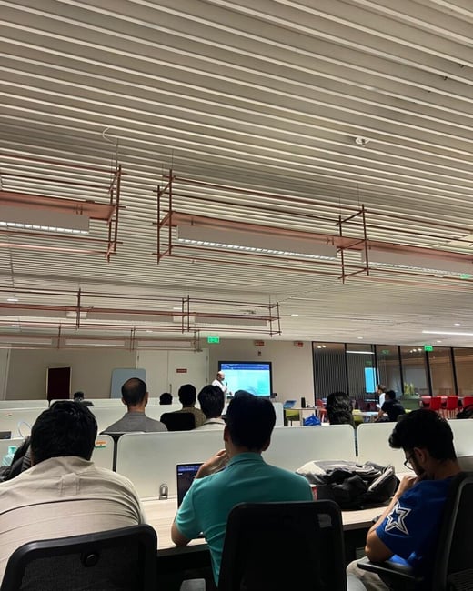 A group of people are seated in a modern office space, attending a presentation given by a speaker at the front of the room.