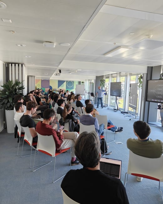 A man is giving a presentation to a group of people seated in a modern, brightly lit office space.