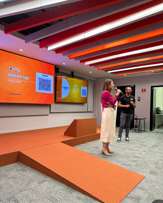A woman in a magenta top and cream skirt speaks into a microphone on a stage during an 'AI Tinkerers' meetup event, with a man standing nearby and large screens displaying event details.