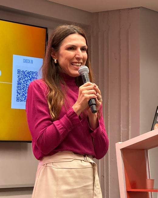 A woman with long brown hair, wearing a magenta blouse and a beige skirt, is smiling and speaking into a microphone during a presentation.