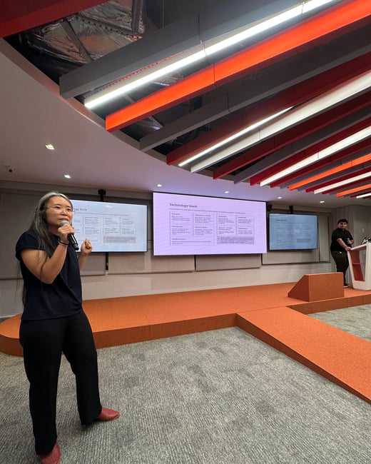 A woman is giving a technical presentation about a technology stack in a modern room with orange accents and multiple screens.