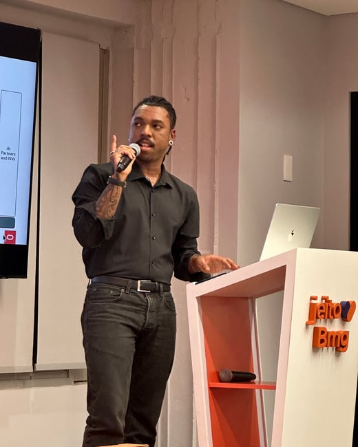 A man in a black shirt and jeans is giving a presentation at a podium, holding a microphone and gesturing while standing next to a laptop and a presentation screen.