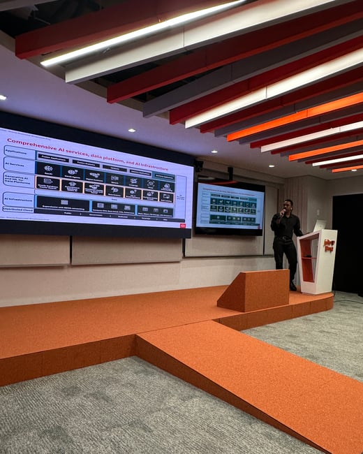 A man stands on an orange stage giving a presentation about AI services, with a large screen behind him displaying a technical diagram.