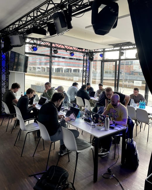 A group of people are working on laptops at long white tables in a modern, well-lit indoor space. The room features large windows with a city view and a professional lighting rig on the ceiling.