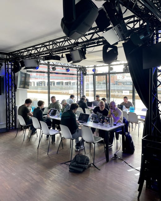 A group of people are working on laptops at long tables inside a modern studio or coworking space equipped with professional lighting rigs. Large windows in the background offer a view of an urban waterfront area.