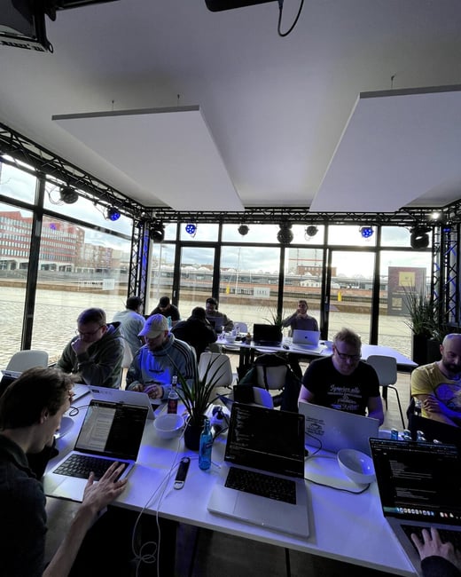 A group of people are sitting at a long white table, deeply focused on their laptops in a modern room with large glass windows overlooking a waterfront.