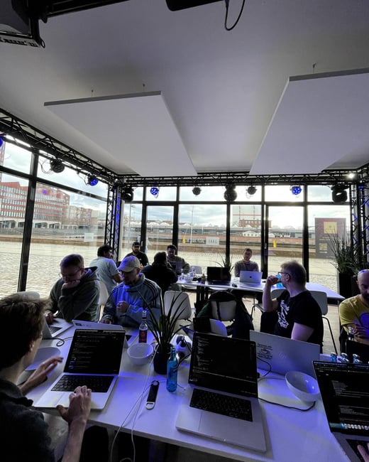 A group of people are gathered in a modern, glass-walled room, focused on their laptops at long tables, likely participating in a hackathon or collaborative work session.