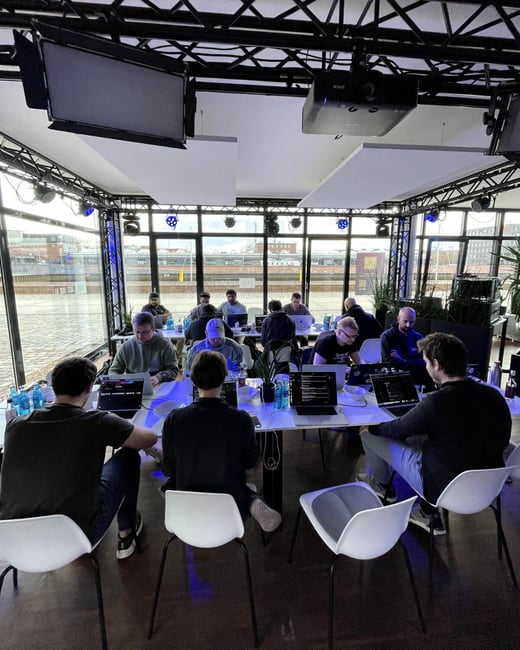 A group of people are seated at long white tables in a modern, glass-walled room, focused on their laptops during what appears to be a coding event or hackathon.