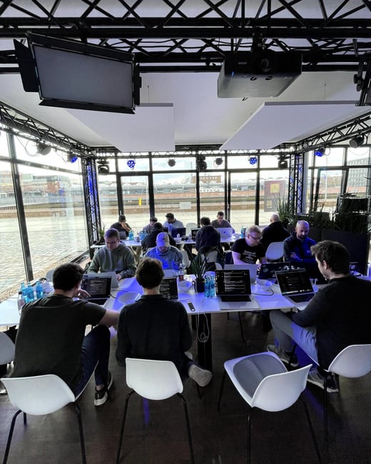 A group of individuals are gathered in a modern, glass-walled room, intensely focused on their laptops at long tables.
