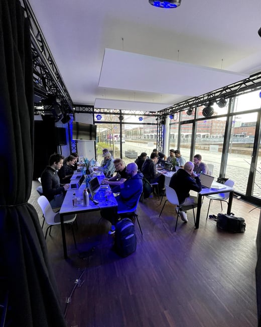 A group of people are seated at long tables in a modern indoor space with large windows, focused on their laptops during a collective working or coding session.