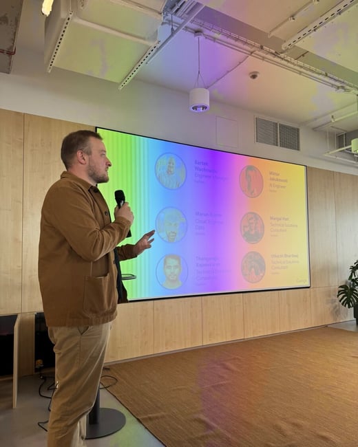 A man stands in profile, holding a microphone and gesturing during a presentation in a modern office space. Behind him is a large screen displaying a colorful grid of team member profiles with names and job titles.