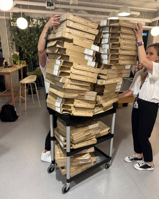 A large stack of Domino's pizza boxes on a black rolling cart being handled by two people in an office-like setting.