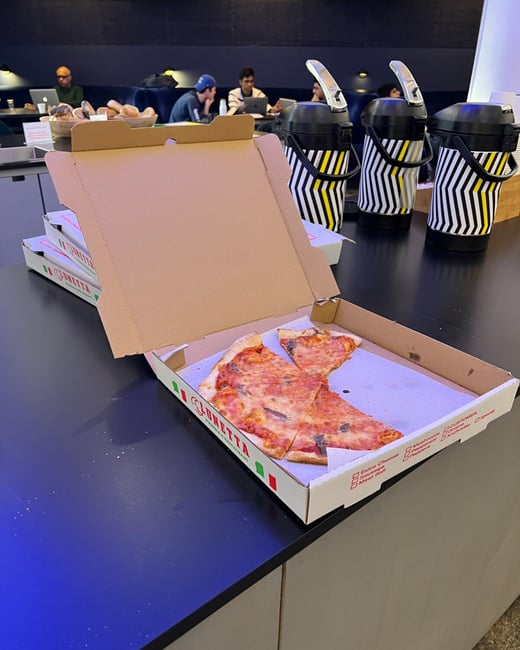A candid photograph shows an open pizza box with a few remaining slices on a dark countertop, suggesting a break or meal during a working session in a modern, dimly lit office space.