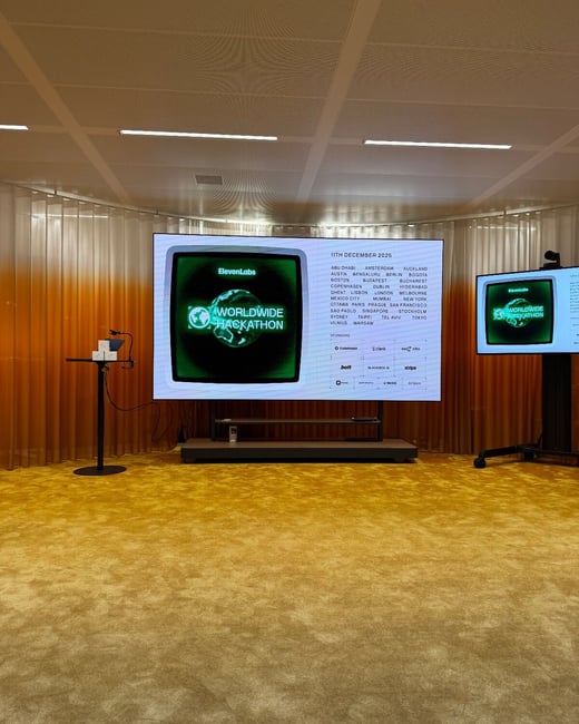 A photograph of an empty, modern event space featuring a large central screen displaying details for the 'WORLDWIDE HACKATHON' organized by ElevenLabs, set against a backdrop of orange gradient curtains and gold carpet.