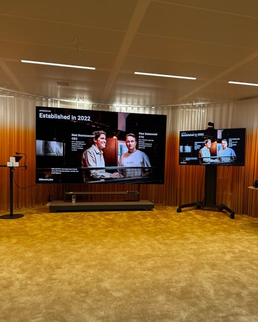 A photograph showing an empty modern presentation room featuring two large screens displaying a slide about the founders (Mati Staniszewski and Piotr Dabkowski) of a company established in 2022, set against a backdrop of orange gradient curtains and a bright yellow carpet.