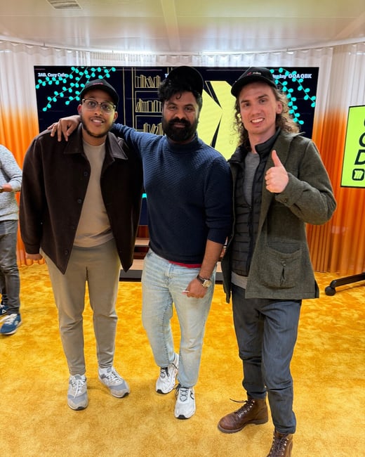A group portrait of three men standing together and smiling in an indoor setting, characterized by a bright yellow carpet and a large digital screen backdrop displaying abstract graphics and text.